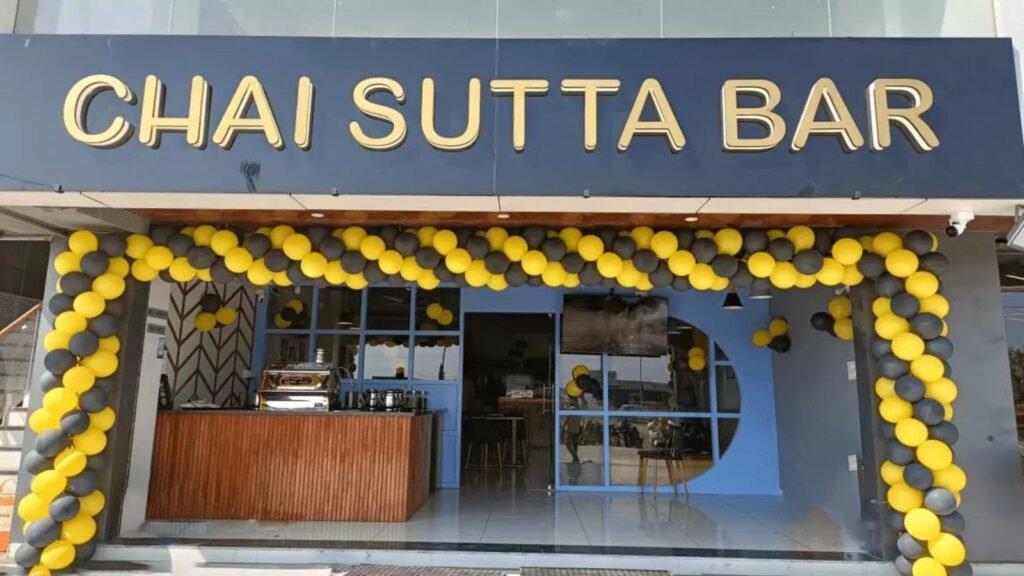 A storefront with a "Chai Sutta Bar" sign above the entrance. The entrance is decorated with yellow and black balloons. There is a coffee machine and seating area visible inside the shop you will Get the Chai Sutta Bar Menu With Prices Jammu