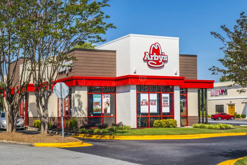 An Arby's fast-food restaurant with a white and brown building, a red roof, and a large Arby's logo. The restaurant is surrounded by trees and a parking lot. Is Arby's Open On Labor Day