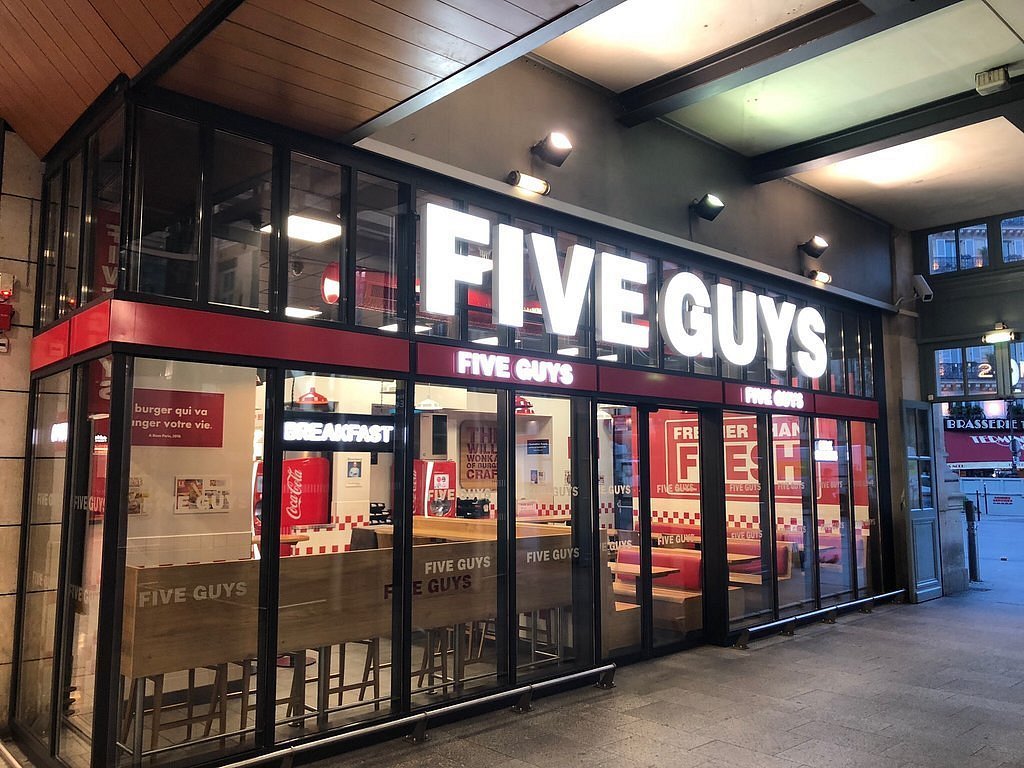 A brightly lit Five Guys restaurant with large windows and a red and white sign. The restaurant has a modern and inviting atmosphere with comfortable seating and a variety of menu options also showcasing Is Five Guys open on Labor Day