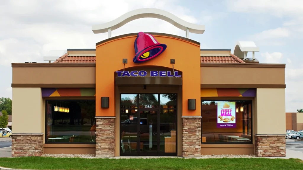 A brightly lit Taco Bell restaurant with a drive-thru lane. The restaurant is open and welcoming, with customers visible inside. Is Taco Bell open on Labor Day? Find out here!