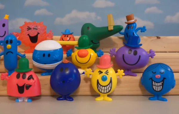 McDonald's Under 3 Toys A group of Mr. Men and Little Miss toys standing on a wooden surface. The toys are brightly colored and have simple, expressive faces. From left to right, we see Mr. Jelly, Mr. Messy, Mr. Small, Mr. Crocodile, Mr. Top Hat, Mr. Bump, Mr. Happy, Mr. Tickle, Mr. Greedy, Mr. Tall, Mr. Mean and Mr. Cheerful.